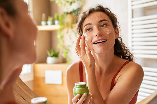 Woman looking in mirror as she applies cream from a jar on her face.