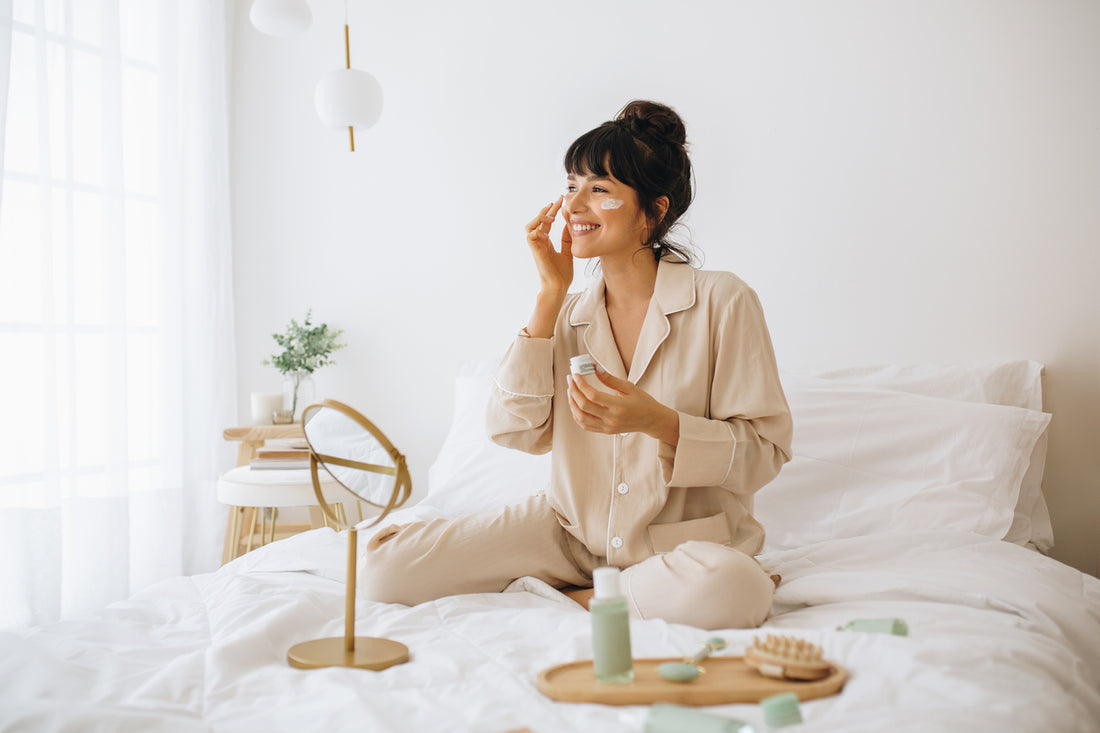 A brunette woman sitting on her bed in pajamas applying cream to her face as part of her skin care routine.